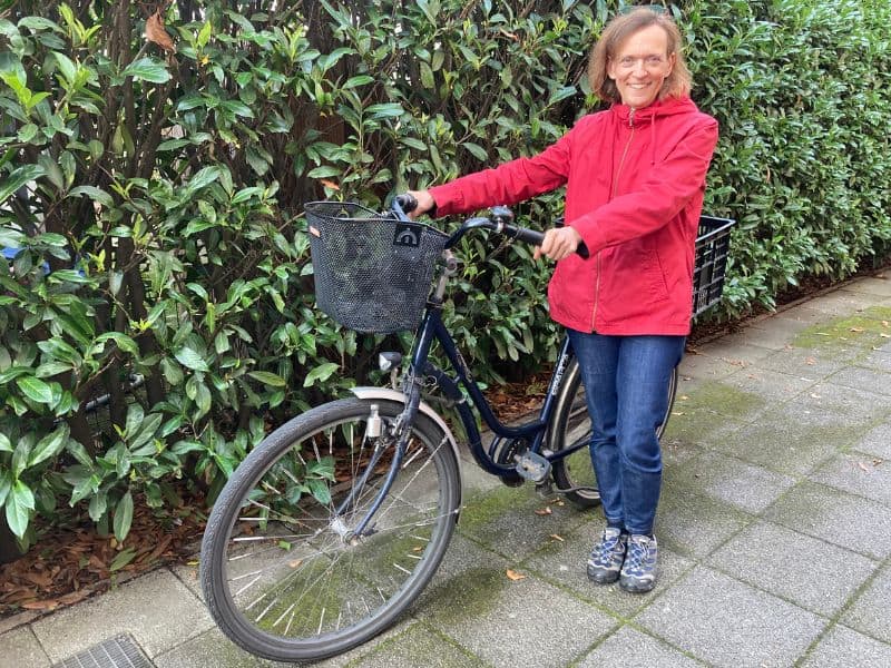 Frau in roter Jacke und Jeans steht neben einem blauen Fahrrad mit Korb, vor einer dichten Kirschlorbeerhecke.